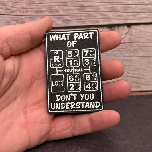 A hand holding a black rectangular patch with white text and car gear symbols. The patch reads, 'WHAT PART OF DON'T YOU UNDERSTAND.
