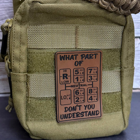 An olive green tactical pouch with a brown woven strap and a central tan patch that reads 'WHAT PART OF DON'T YOU UNDERSTAND' with a periodic table-style graphic, shown against a gray wood plank background.