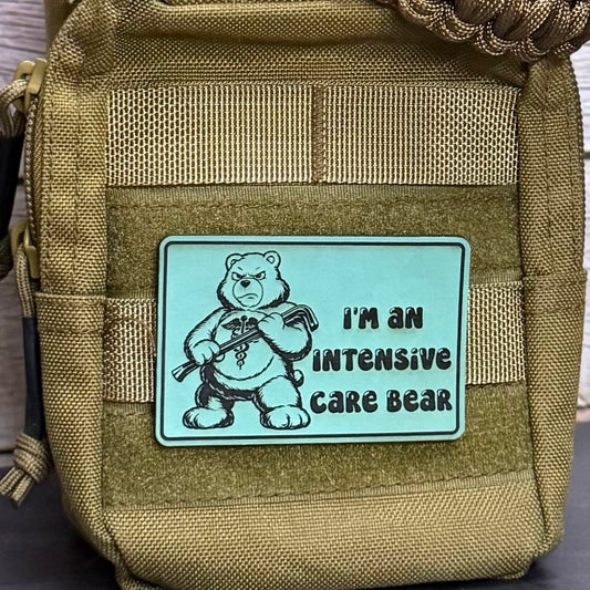 A close-up of a tactical pouch in coyote brown webbed fabric, featuring a rectangular light blue patch with a cartoon grumpy bear and the text 'I'M AN INTENSIVE CARE BEAR'.