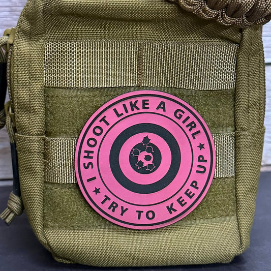 An olive green tactical pouch with a pink and black circular patch that reads 'I SHOOT LIKE A GIRL TRY TO KEEP UP' around a target graphic, shown against a light wood background.