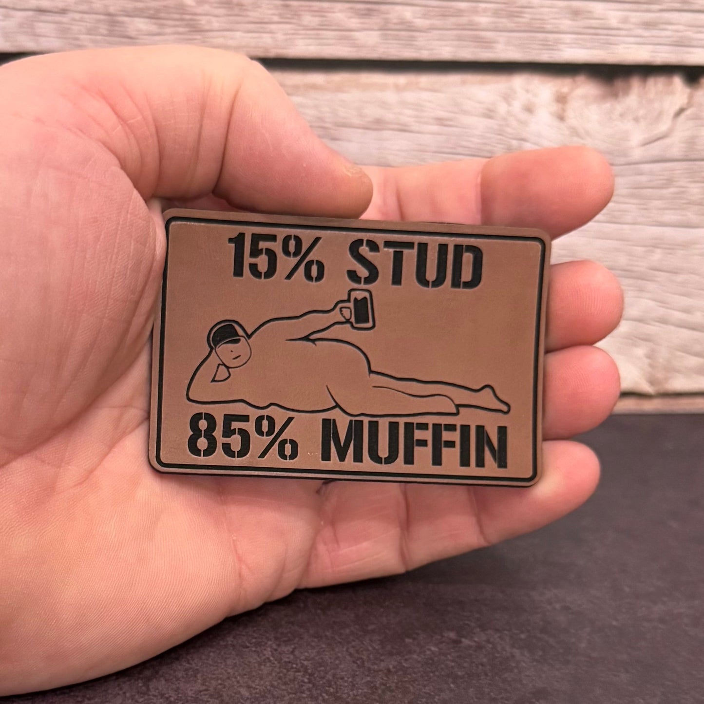 A hand holding a rectangular metal pin with a humorous slogan reading '15% STUD 85% MUFFIN' and an illustration of a person lying down taking a selfie, set against a rustic wood background.