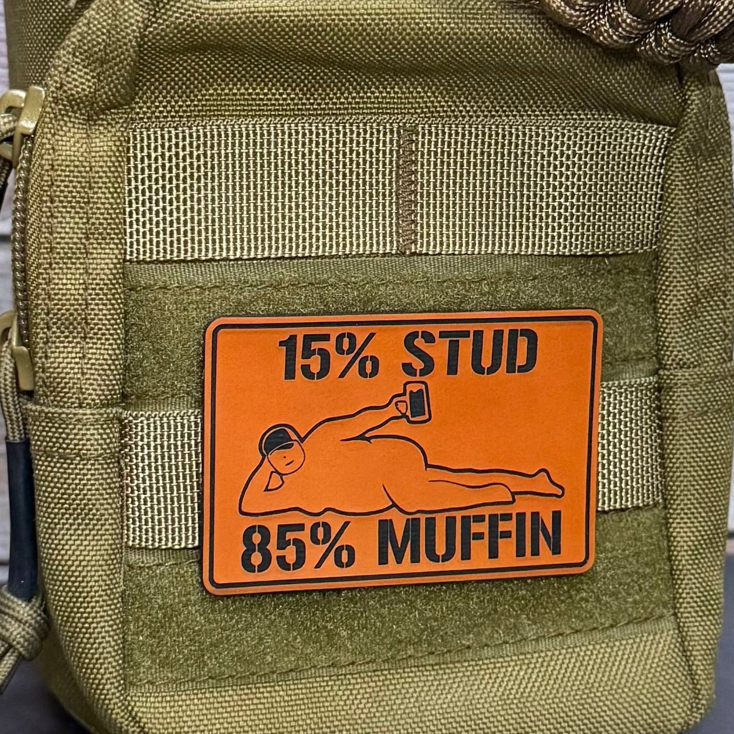 An olive green tactical pouch with a prominent orange rectangular patch that reads '15% STUD 85% MUFFIN' with a graphic of a person lying down holding a mug. The pouch has MOLLE webbing and a zipper on the left side.