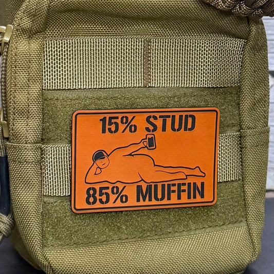 An olive green tactical pouch with a prominent orange rectangular patch that reads '15% STUD 85% MUFFIN' with a graphic of a person lying down holding a mug. The pouch has MOLLE webbing and a zipper on the left side.