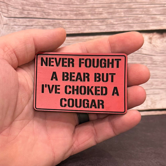 A hand holding a rectangular red novelty patch with black text that reads 'NEVER FOUGHT A BEAR BUT I'VE CHOKED A COUGAR' against a rustic wood plank background.