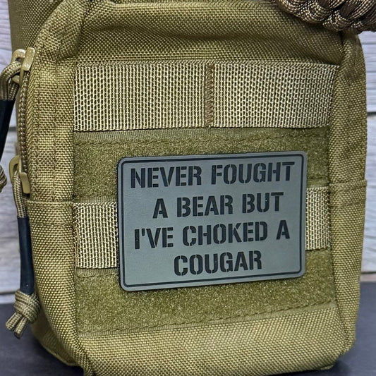 An olive green tactical pouch with a gray rectangular patch that reads 'NEVER FOUGHT A BEAR BUT I'VE CHOKED A COUGAR' in black text, shown against a gray wood-plank background.