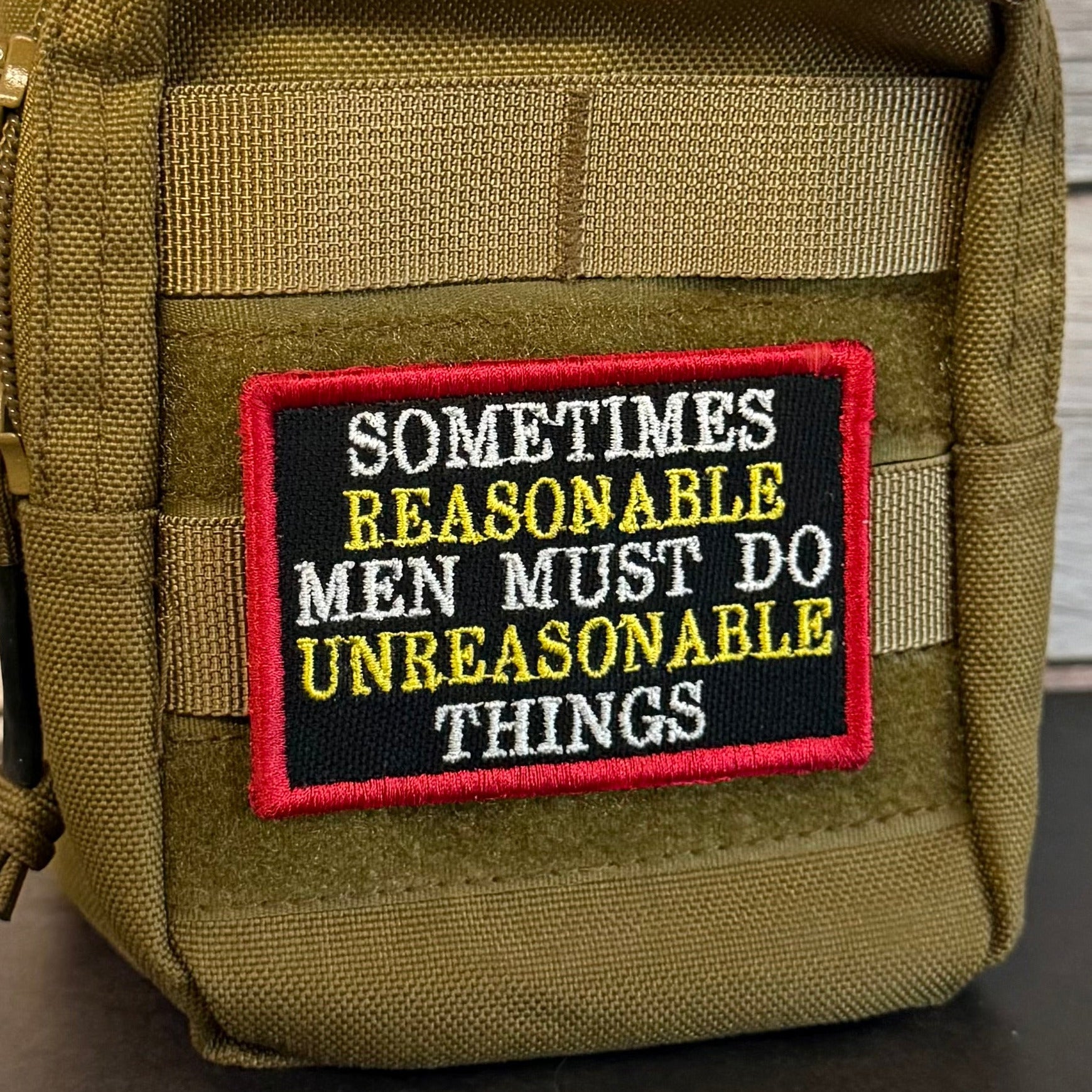 An olive green tactical pouch with a black embroidered patch featuring the text 'SOMETIMES REASONABLE MEN MUST DO UNREASONABLE THINGS' in white and yellow, with a red border.