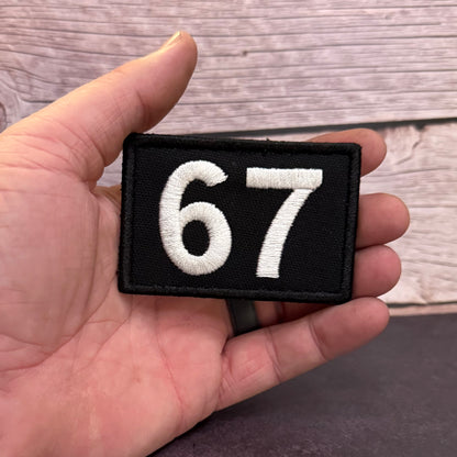 A hand holding a small rectangular black patch with the white embroidered numbers 67, against a rustic wood plank background.
