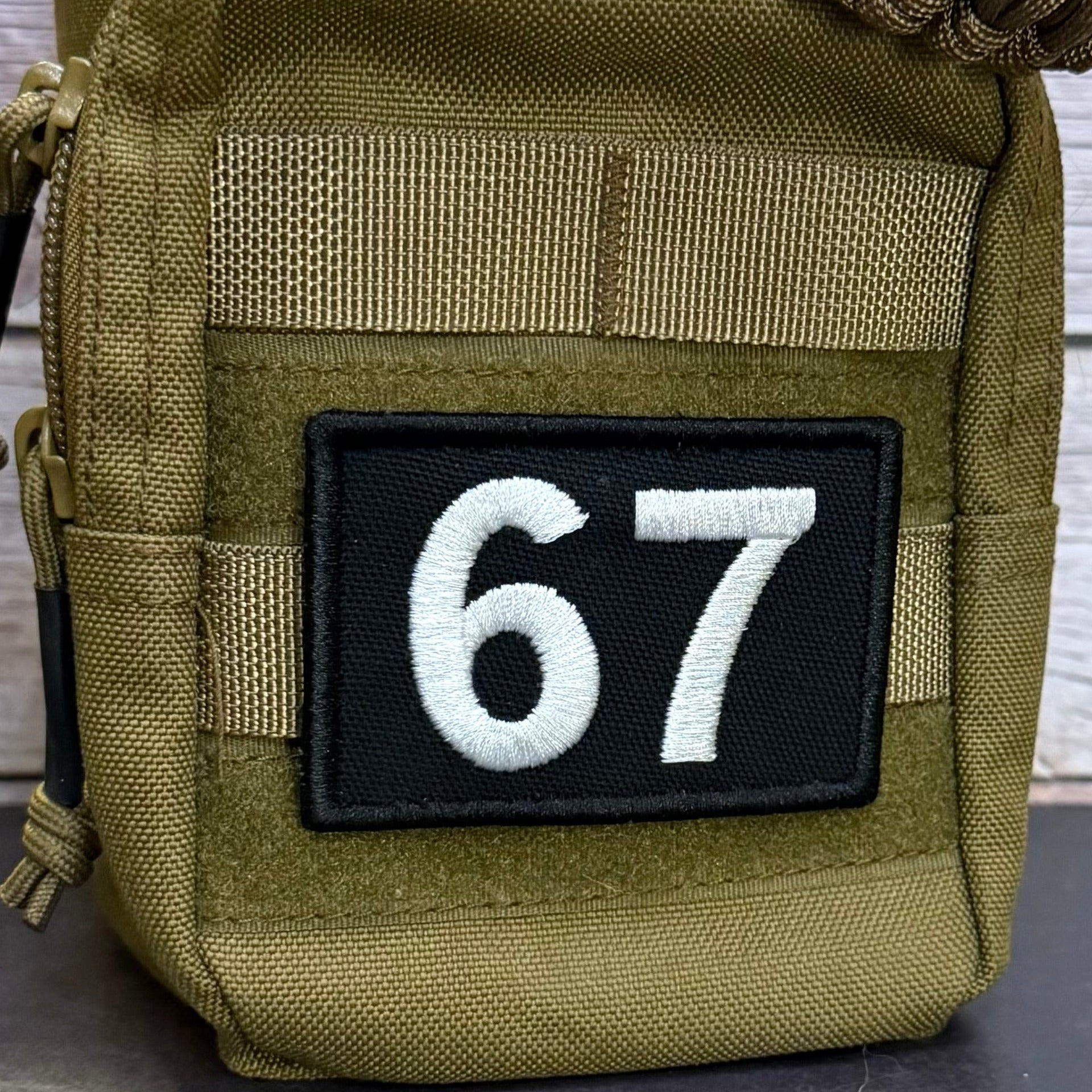 Close-up of a tactical pouch in olive drab green webbing with a black rectangular patch featuring the white embroidered numbers 67, shown against a light wood plank background.