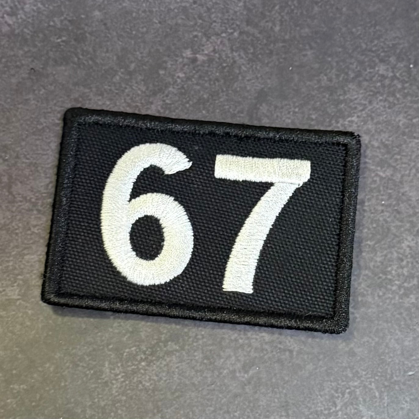 A rectangular black embroidered patch with the white numbers '67' on a textured gray background.