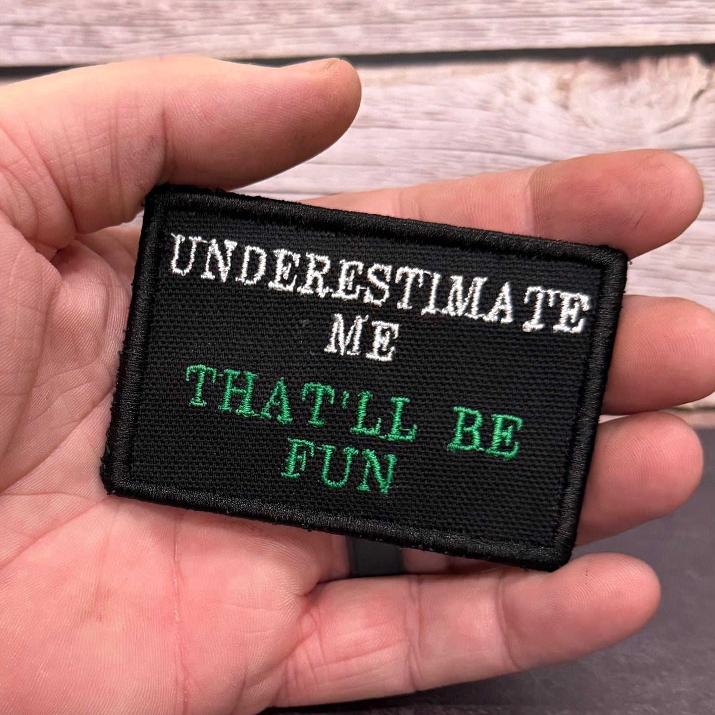 A hand holding a rectangular black embroidered patch with the text 'UNDERESTIMATE ME THAT'LL BE FUN' in white and green lettering, set against a rustic wood plank background.
