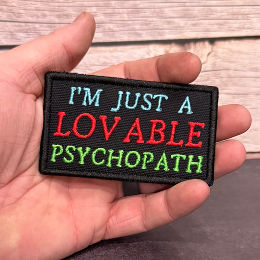 A hand holding a rectangular embroidered patch that reads 'I'M JUST A LOVABLE PSYCHOPATH' in light blue, red, and green lettering on a black background, with a wood plank surface behind it.