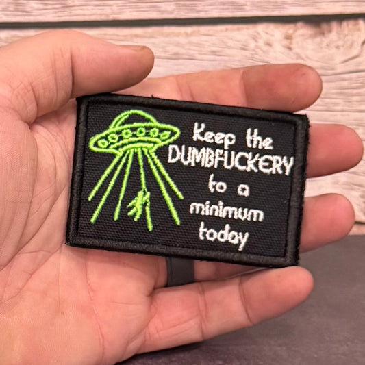 A hand holding a rectangular black embroidered patch with a green UFO graphic and white text reading 'Keep the DUMBFUCKERY to a minimum today', set against a rustic wood plank background.