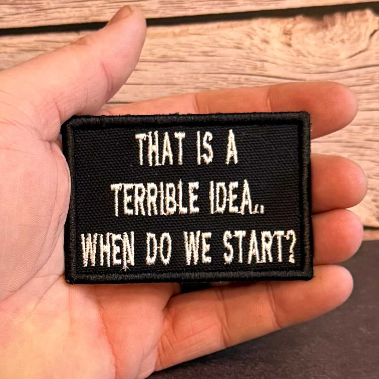 A hand holding a rectangular black embroidered patch with white text that reads, 'THAT IS A TERRIBLE IDEA.