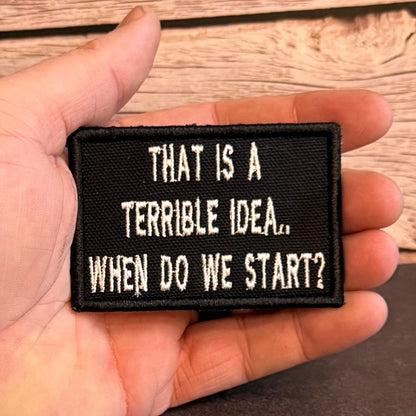 A hand holding a rectangular black embroidered patch with white text that reads, 'THAT IS A TERRIBLE IDEA.