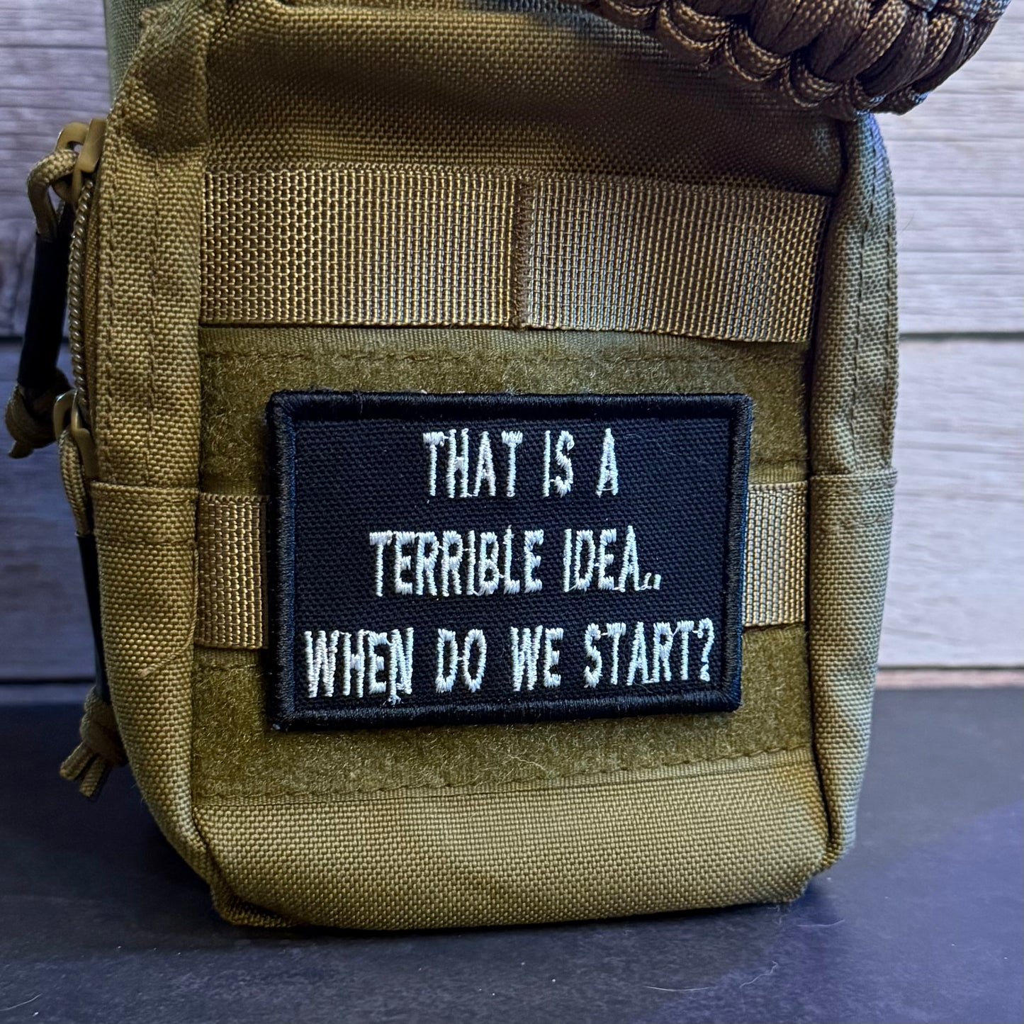 A close-up of a tactical pouch in olive drab nylon with a black rectangular patch featuring the white embroidered text 'THAT IS A TERRIBLE IDEA.