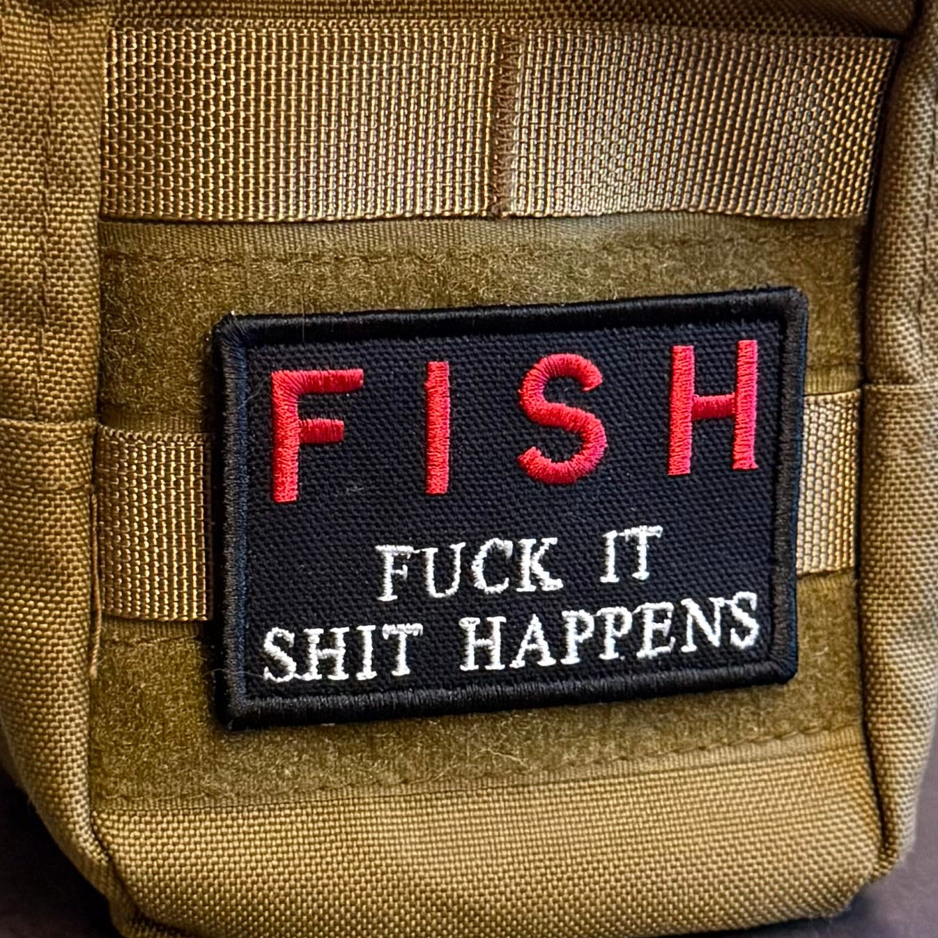 A close-up of a tan tactical pouch featuring a black rectangular patch with the word 'FISH' in red and the phrase 'FUCK IT SHIT HAPPENS' in white text.