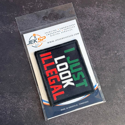 A rectangular embroidered patch in clear plastic packaging, featuring the text 'I JUST LOOK ILLEGAL' in red, white, and green on a black background. The packaging card shows the brand 'Jeksp Service'.