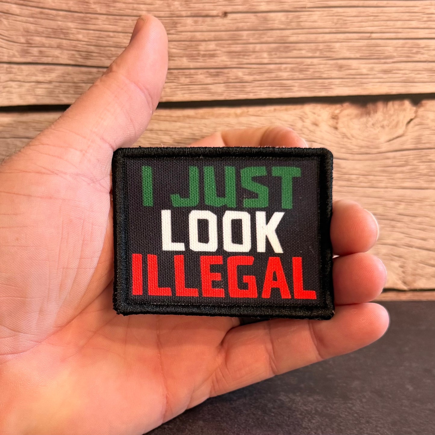A rectangular embroidered patch held in a hand, featuring the text 'I JUST LOOK ILLEGAL' in green, white, and red lettering on a black background, with a wood plank surface behind it.