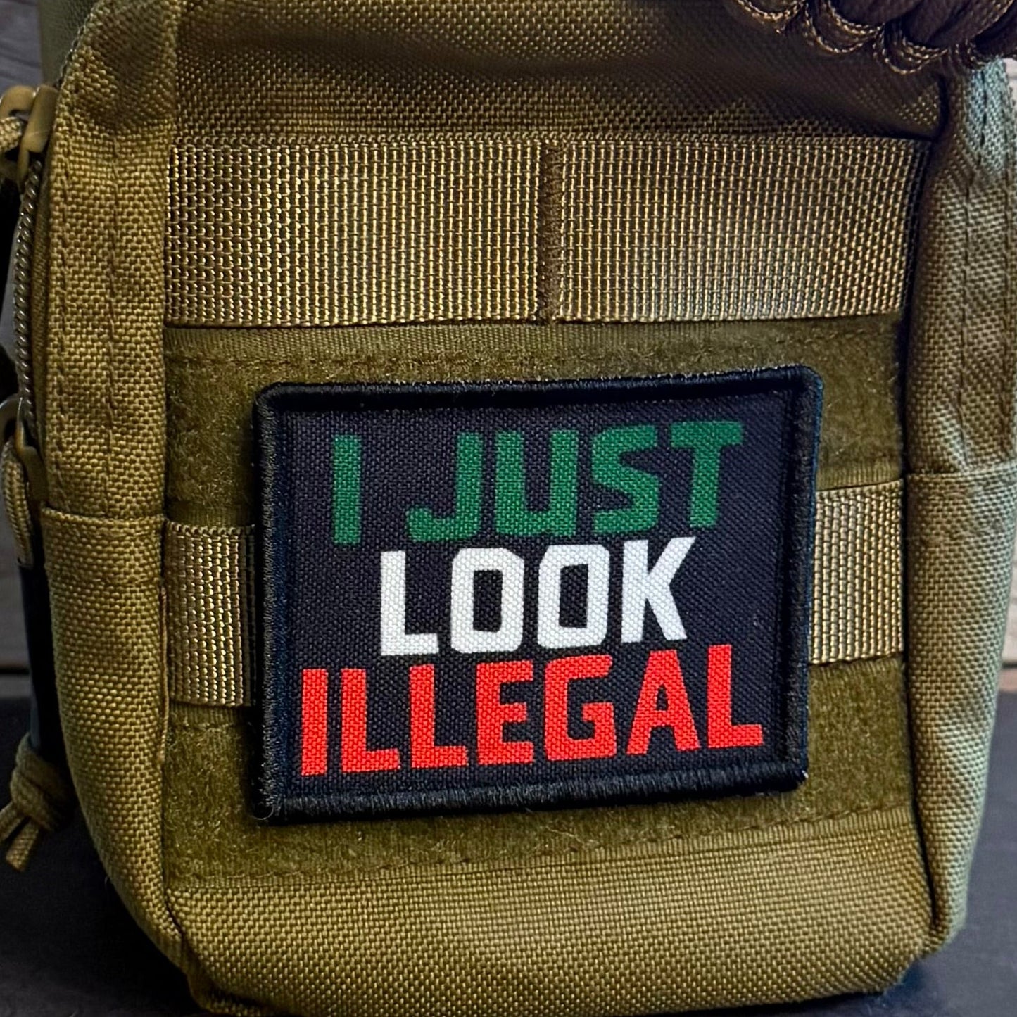 A close-up of a tactical pouch in olive drab green nylon with a black rectangular patch that reads 'I JUST LOOK ILLEGAL' in green, white, and red lettering. A brown paracord strap is threaded through the top, and the pouch is set against a rustic wood plank background.