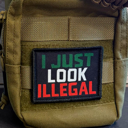 A close-up of a tactical pouch in olive drab green nylon with a black rectangular patch that reads 'I JUST LOOK ILLEGAL' in green, white, and red lettering. A brown paracord strap is threaded through the top, and the pouch is set against a rustic wood plank background.
