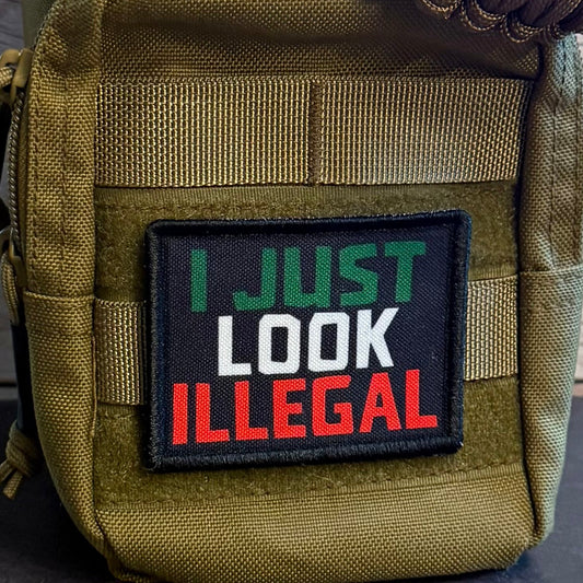 A close-up of a tactical pouch in olive drab green nylon with a black rectangular patch that reads 'I JUST LOOK ILLEGAL' in green, white, and red lettering. A brown paracord strap is threaded through the top, and the pouch is set against a rustic wood plank background.