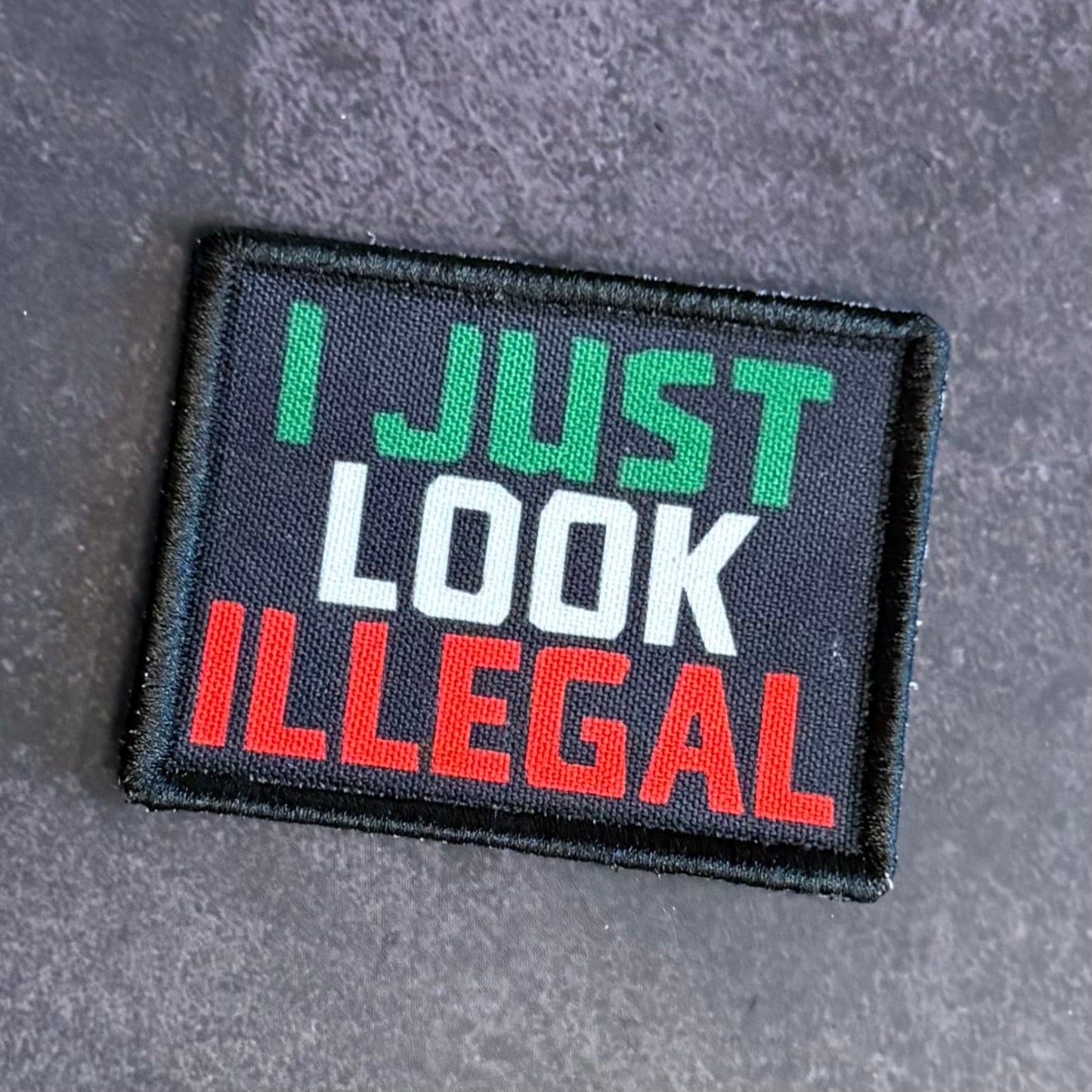 A rectangular embroidered patch with the phrase 'I JUST LOOK ILLEGAL' in green, white, and red lettering on a black background, placed on a textured gray surface.