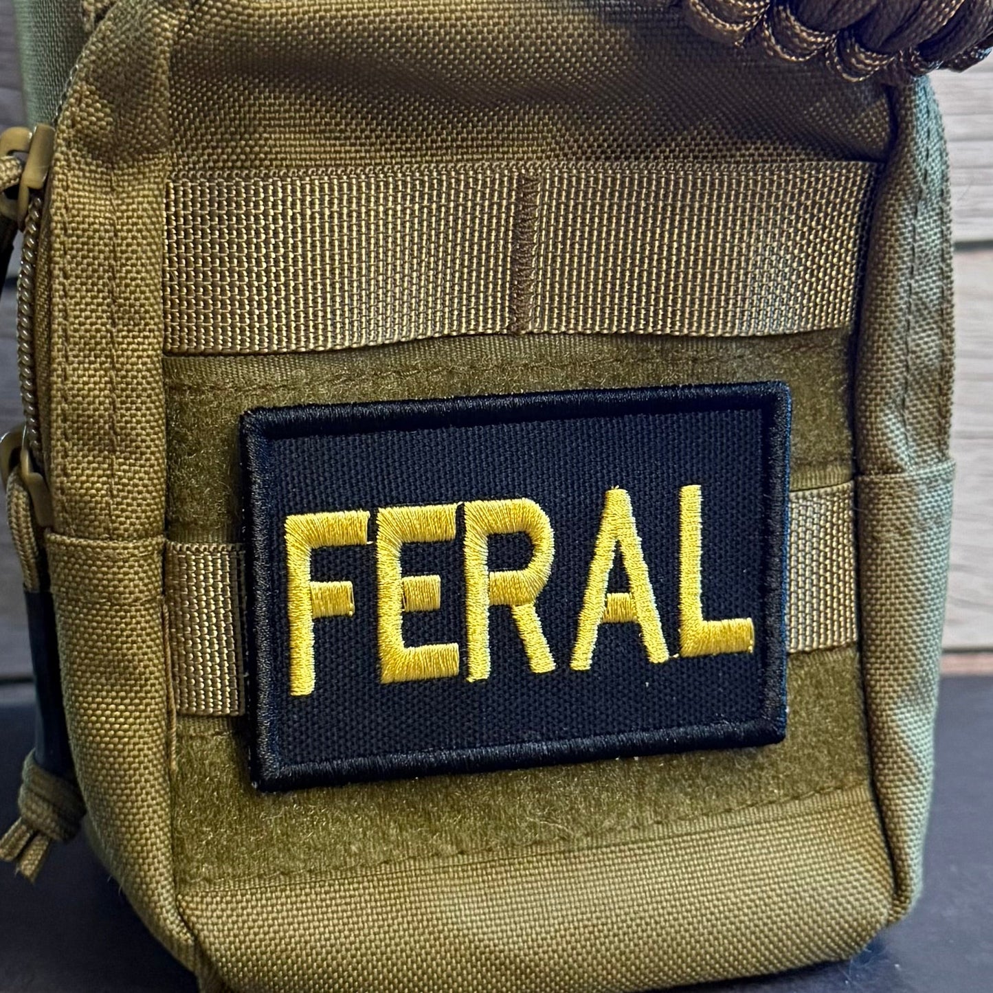 A close-up of a tactical pouch in olive drab green nylon with a black rectangular patch featuring the word 'FERAL' in yellow embroidered letters. A brown paracord strap is visible at the top, and the pouch is photographed against a dark surface with a wood plank background.