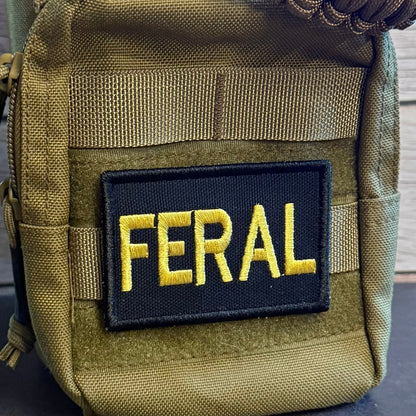 A close-up of a tactical pouch in olive drab green nylon with a black rectangular patch featuring the word 'FERAL' in yellow embroidered letters. A brown paracord strap is visible at the top, and the pouch is photographed against a dark surface with a wood plank background.