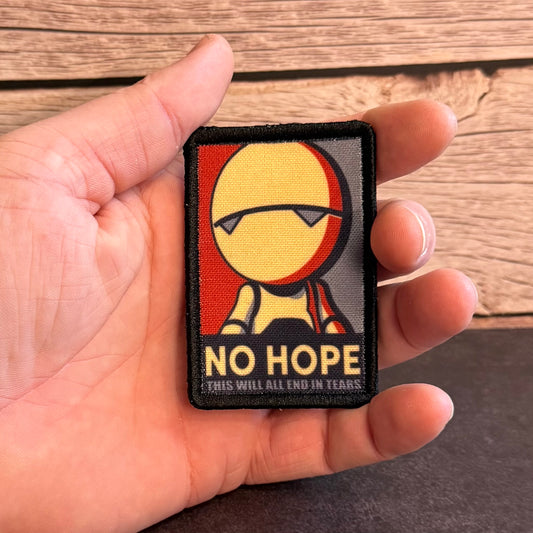 A hand holding a rectangular embroidered patch featuring a stylized yellow face with dark sunglasses, red and gray side panels, and the text 'NO HOPE' and 'THIS WILL ALL END IN TEARS.' against a wood plank background.