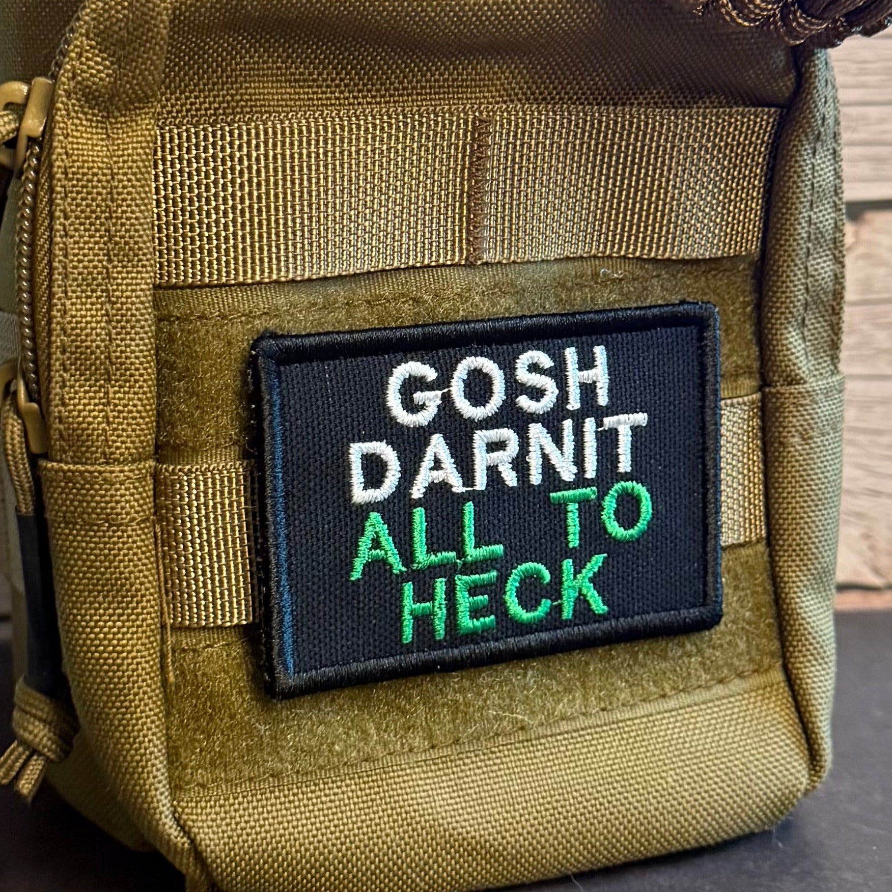 A close-up of a tactical pouch in olive drab nylon with a black rectangular patch that reads 'GOSH DARNIT ALL TO HECK' in white and green embroidery, with a paracord strap visible at the top.