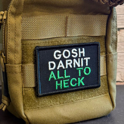 A close-up of a tactical pouch in olive drab nylon with a black rectangular patch that reads 'GOSH DARNIT ALL TO HECK' in white and green embroidery, with a paracord strap visible at the top.