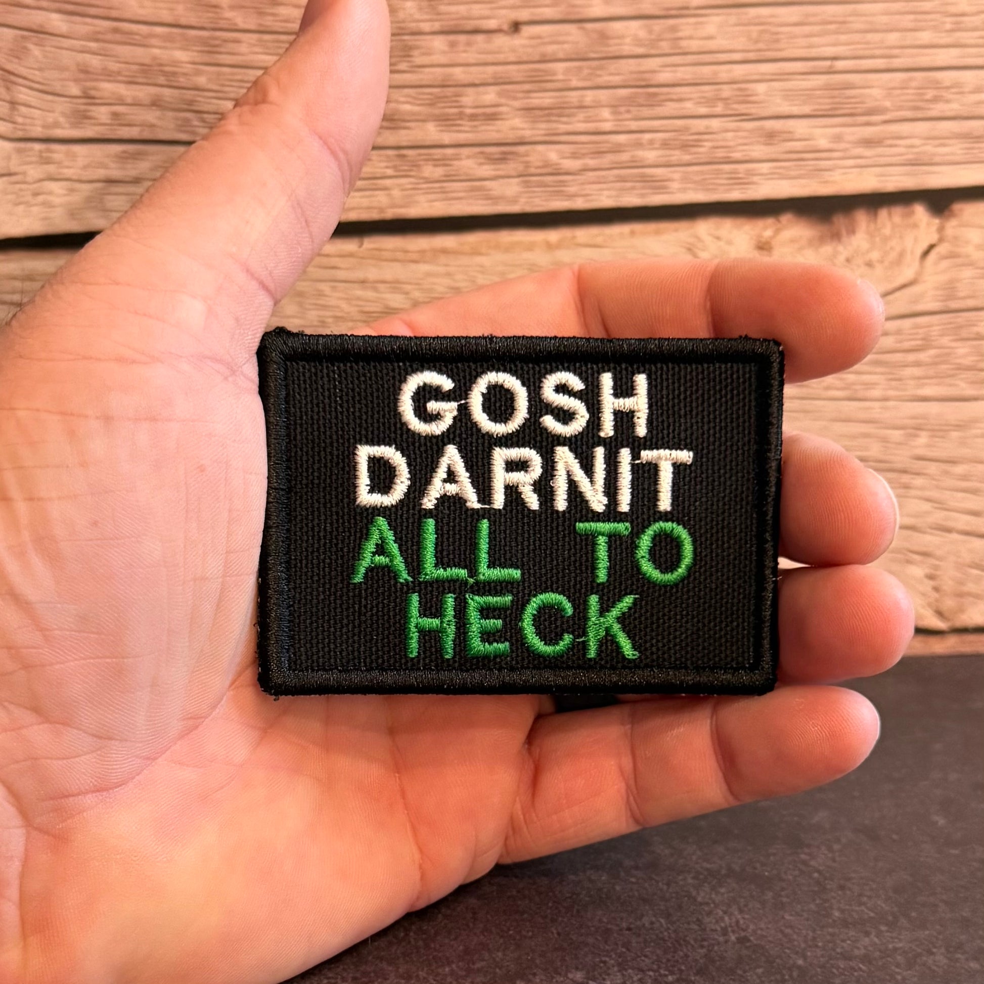 A hand holding a small black embroidered patch with the text 'GOSH DARNIT ALL TO HECK' in white and green lettering, set against a rustic wooden background.