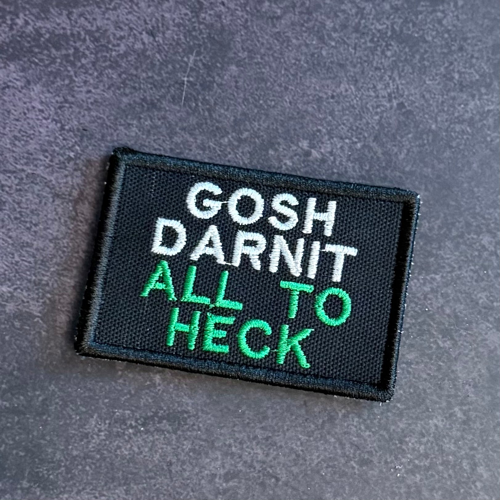 A rectangular embroidered patch with a black background and white and green text reading 'GOSH DARNIT ALL TO HECK', placed on a textured gray surface.