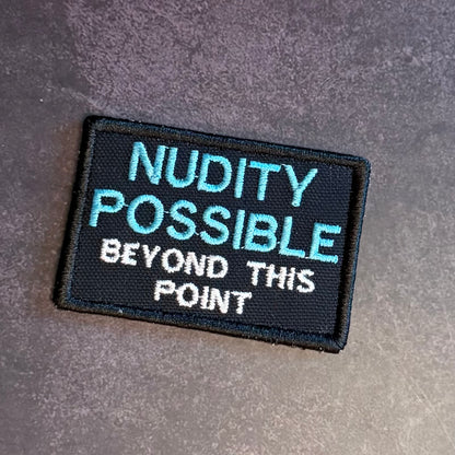 Nude Beach Warning embroidered morale patch: with a black background and the text 'NUDITY POSSIBLE BEYOND THIS POINT' in light blue and white lettering, placed on a textured gray surface.