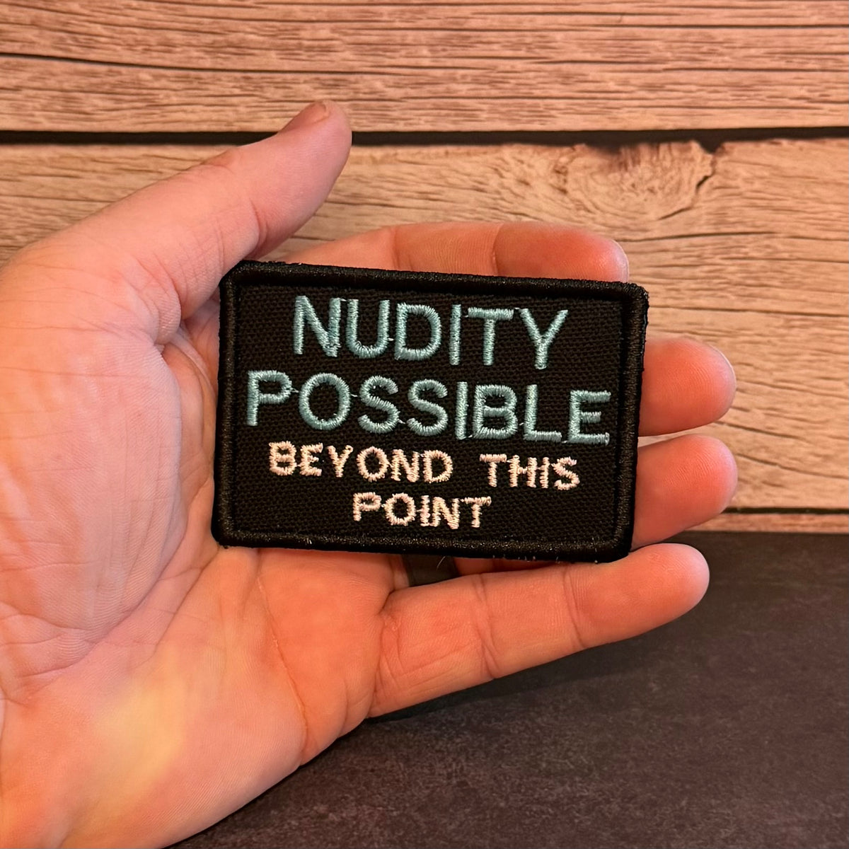 Nude Beach Warning embroidered morale patch: hand holding a rectangular embroidered patch with the text 'NUDITY POSSIBLE BEYOND THIS POINT' in light blue and beige lettering on a black background, set against a wooden surface.