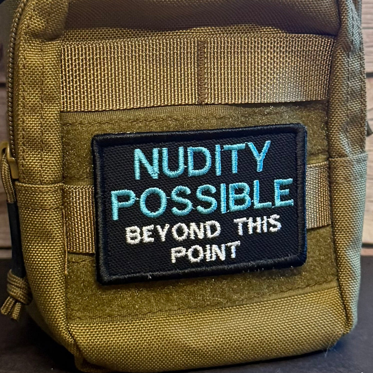Nude Beach Warning embroidered morale patch: tactical pouch in olive drab nylon with a black embroidered patch that reads 'NUDITY POSSIBLE BEYOND THIS POINT' in light blue and white text.