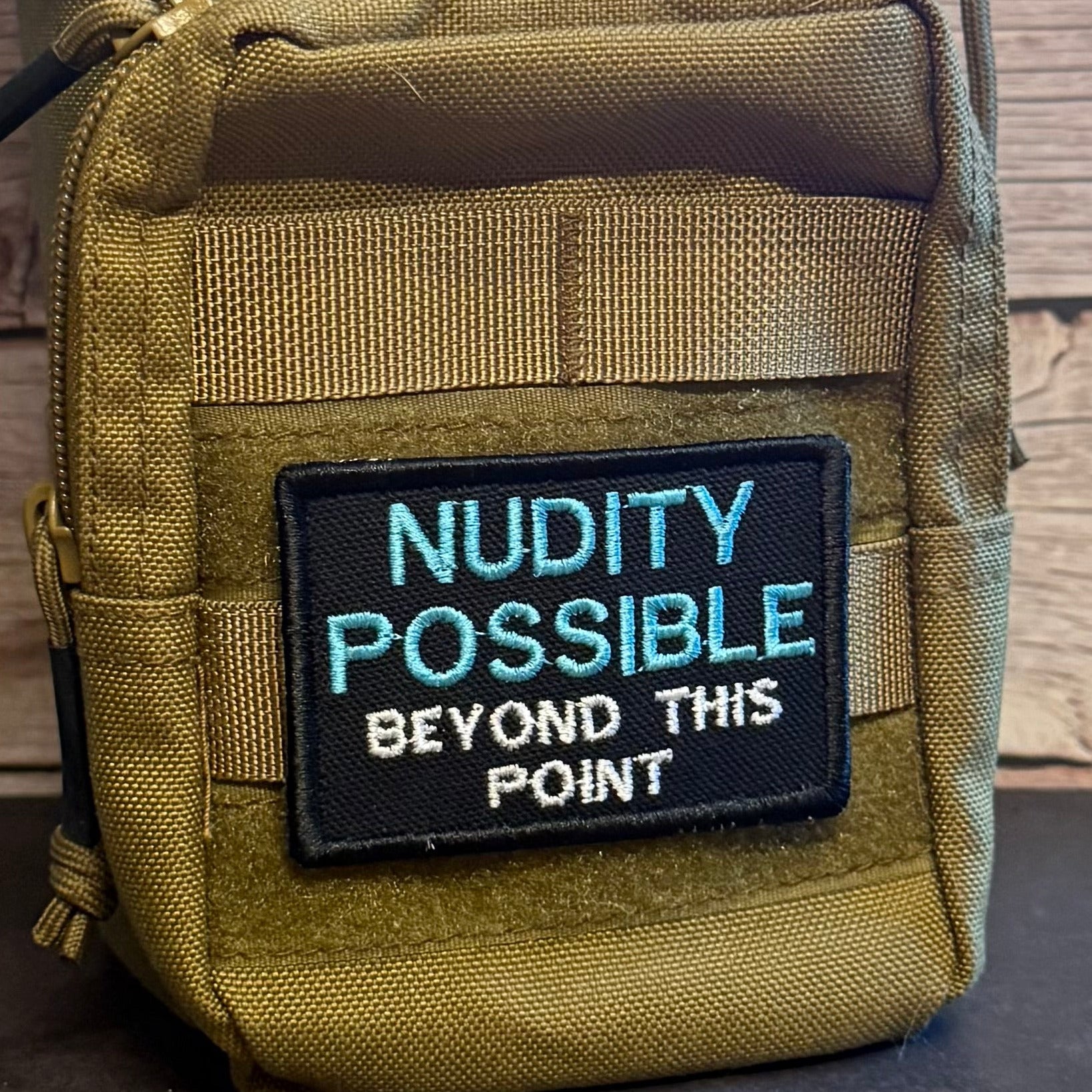 A close-up of a tactical pouch in olive drab nylon with a black embroidered patch that reads 'NUDITY POSSIBLE BEYOND THIS POINT' in light blue and white text.