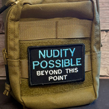 A close-up of a tactical pouch in olive drab nylon with a black embroidered patch that reads 'NUDITY POSSIBLE BEYOND THIS POINT' in light blue and white text.