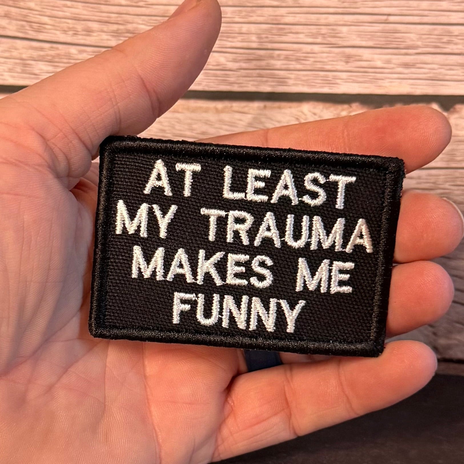 A hand holding a rectangular black embroidered patch with white text that reads 'AT LEAST MY TRAUMA MAKES ME FUNNY' against a rustic wood plank background.