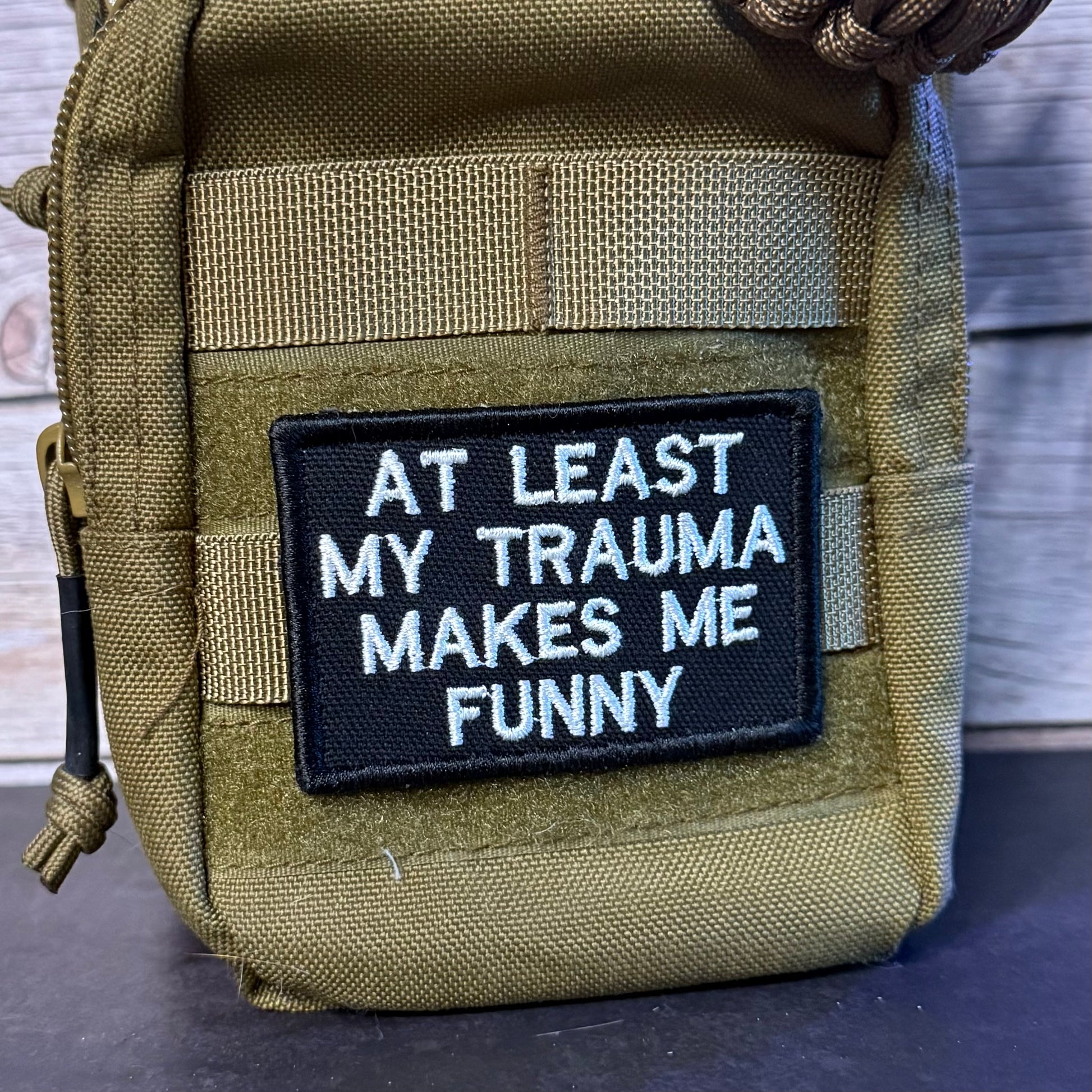 A close-up of a tactical pouch in olive drab green nylon with a black rectangular embroidered patch on the front. The patch features white text that reads 'AT LEAST MY TRAUMA MAKES ME FUNNY'.