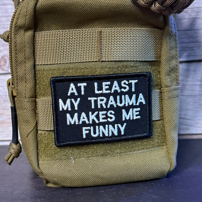 A close-up of a tactical pouch in olive drab green nylon with a black rectangular embroidered patch on the front. The patch features white text that reads 'AT LEAST MY TRAUMA MAKES ME FUNNY'.