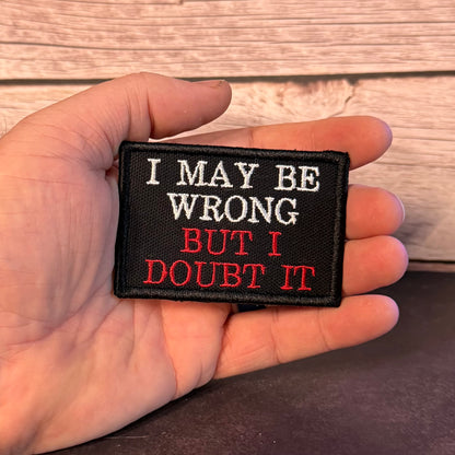 A hand holding a small rectangular embroidered patch with the text 'I MAY BE WRONG BUT I DOUBT IT' in white and red lettering on a black background, set against a rustic wood plank backdrop.