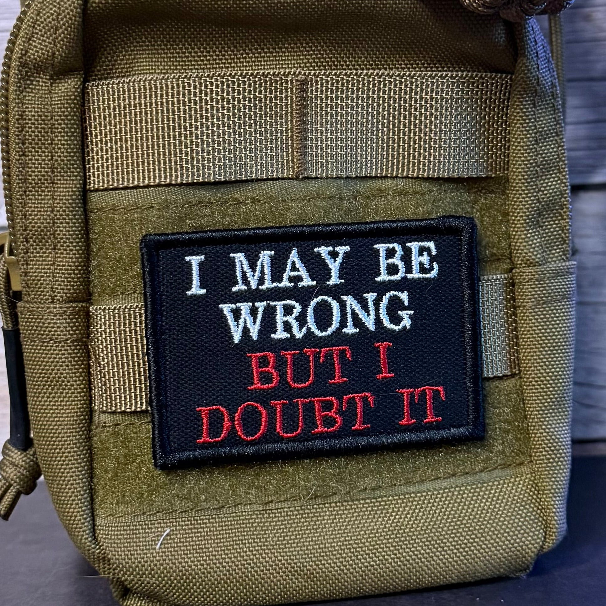 An olive green tactical pouch with a black embroidered patch that reads 'I MAY BE WRONG BUT I DOUBT IT' in white and red text, shown against a dark wood background.