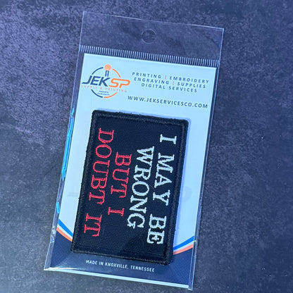 A rectangular black embroidered patch with white and red text reading 'I MAY BE WRONG BUT I DOUBT IT' in a clear plastic sleeve, displayed on a branded card over a dark textured surface.