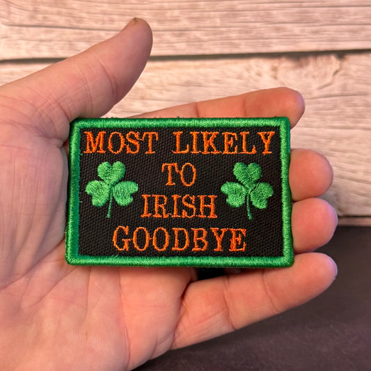 A hand holding a rectangular embroidered patch with the phrase 'MOST LIKELY TO IRISH GOODBYE' in orange text on a black background, with green shamrocks and a green border, against a wood plank background.