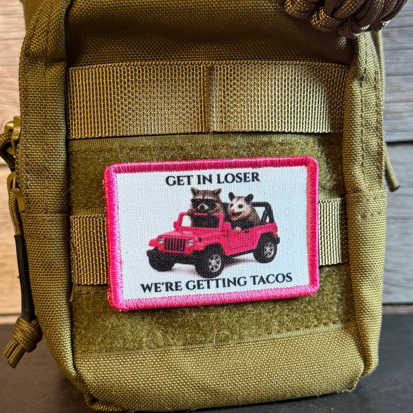An olive green tactical pouch with a rectangular patch featuring a pink border and an illustration of two cartoon animals in a pink jeep with the text 'GET IN LOSER WE'RE GETTING TACOS'.