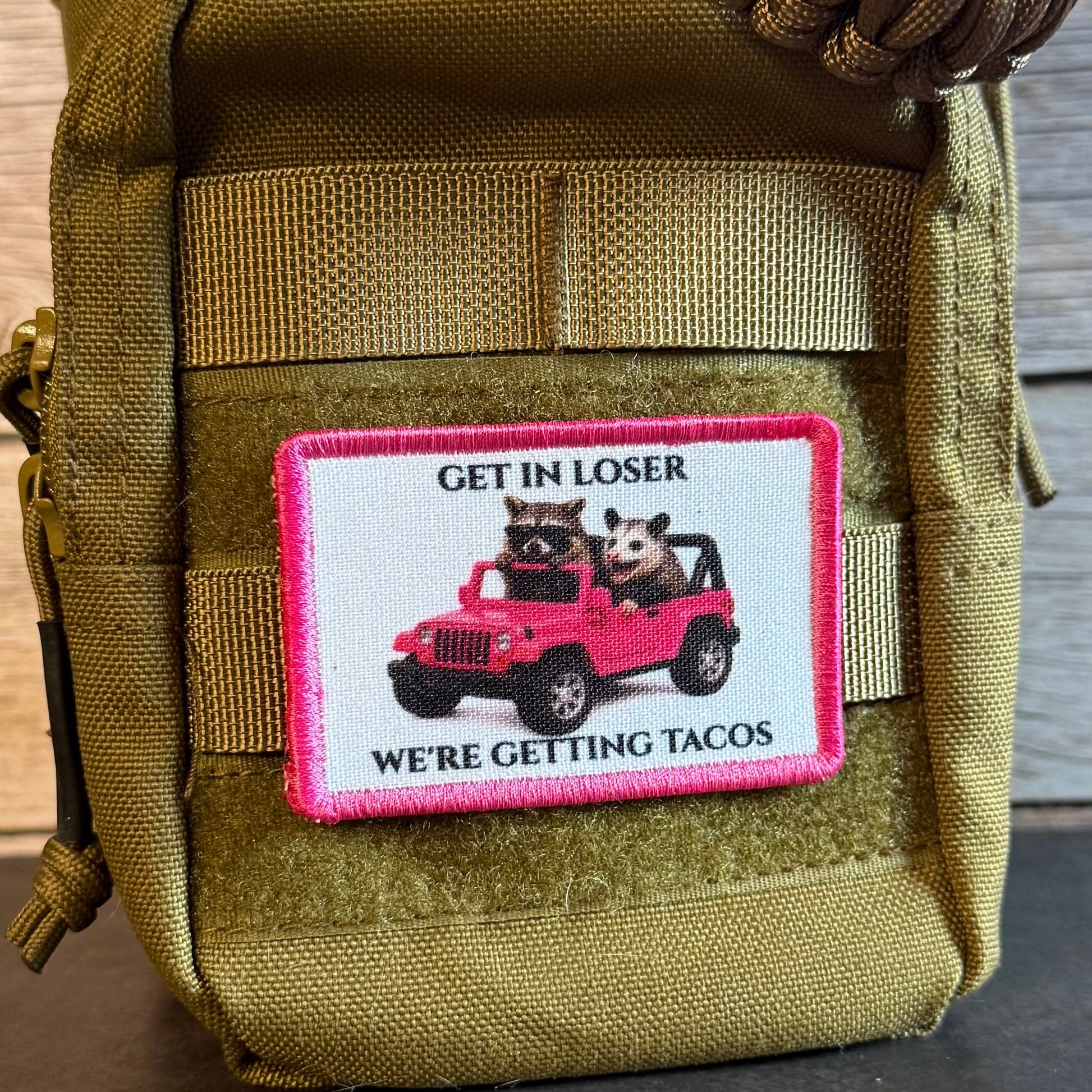 An olive green tactical pouch with a rectangular patch featuring a pink border and an illustration of two cartoon animals in a pink jeep with the text 'GET IN LOSER WE'RE GETTING TACOS'.