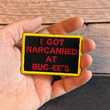 A hand holding a rectangular embroidered patch with a black background, yellow border, and red text that reads 'I GOT NARCANNED AT BUC-EE'S' against a wood plank background.