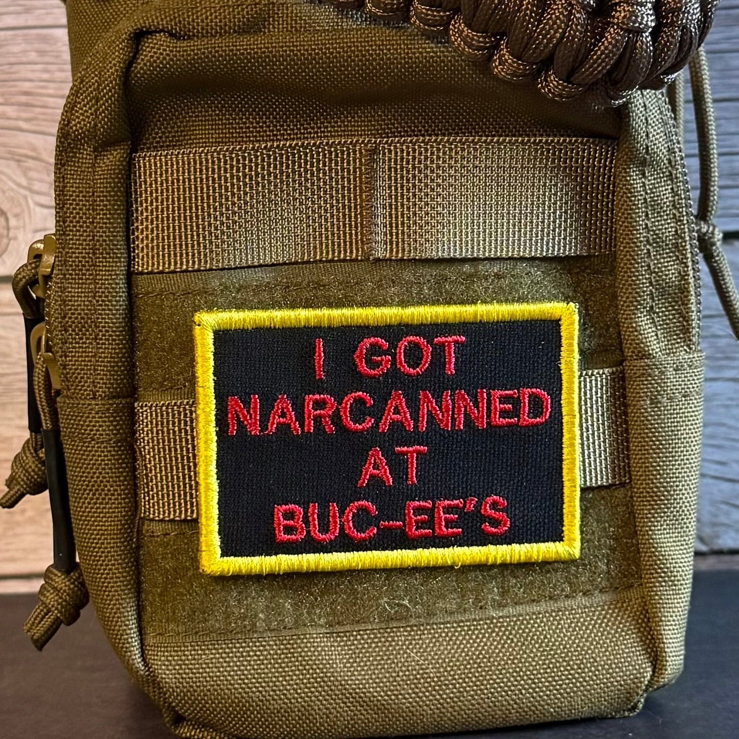 A close-up of a tactical pouch in olive drab nylon with a black embroidered patch featuring red text that reads 'I GOT NARCANED AT BUC-EE'S' and a yellow border. A brown paracord strap is visible at the top, and the pouch is set against a rustic wooden background.