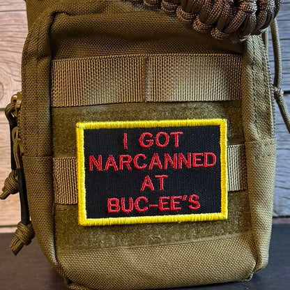 A close-up of a tactical pouch in olive drab nylon with a black embroidered patch featuring red text that reads 'I GOT NARCANED AT BUC-EE'S' and a yellow border. A brown paracord strap is visible at the top, and the pouch is set against a rustic wooden background.